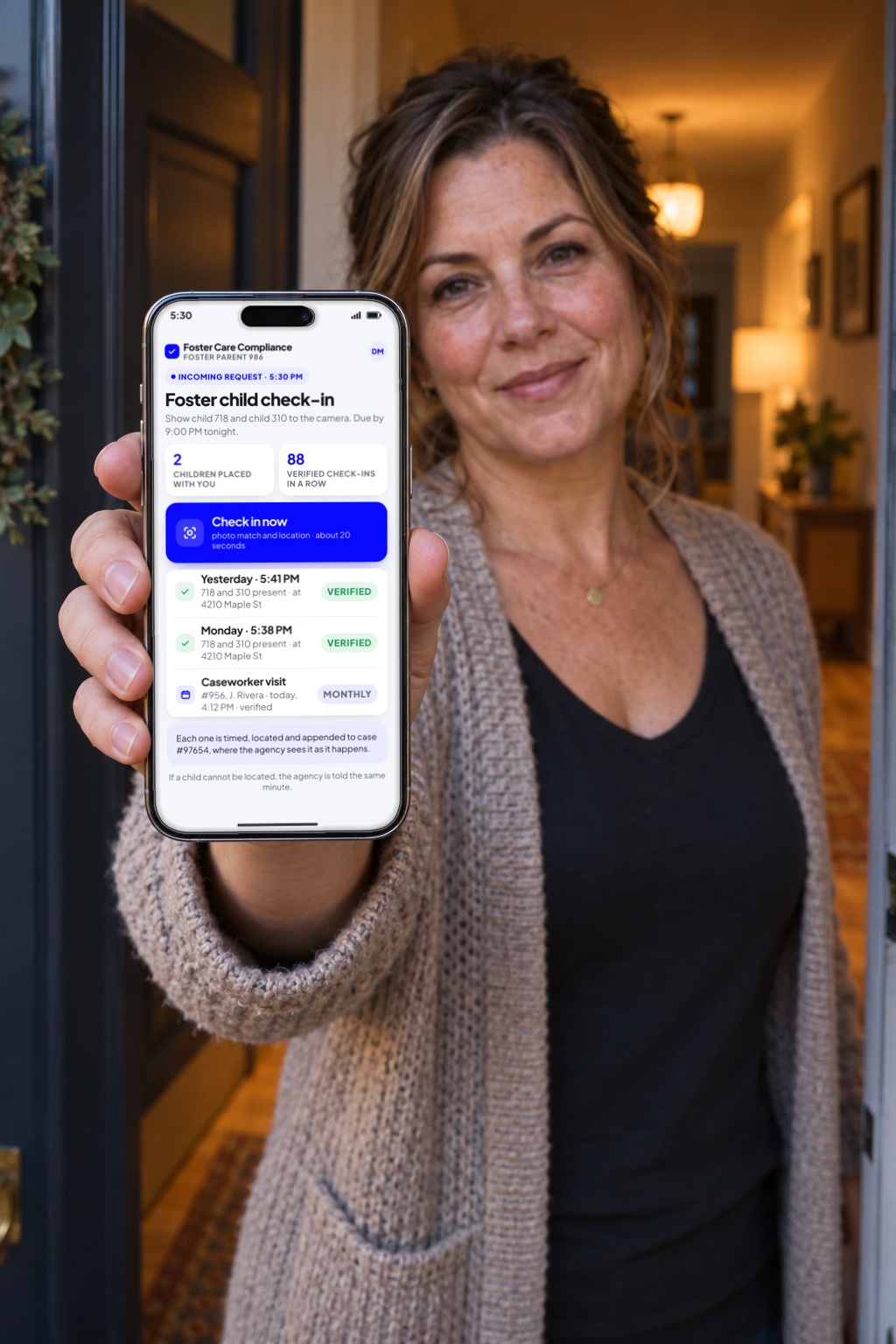 A foster mother in her front doorway holds up her phone showing the daily check-in