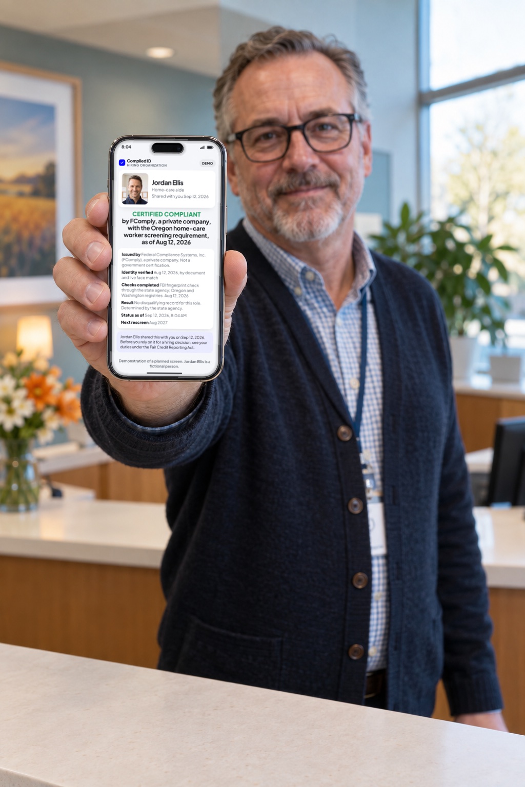 A hospice HR manager at a reception desk holds up his phone showing an illustrative employer's view of a shared Complied ID: CERTIFIED COMPLIANT by FComply, with the photo and name of a fictional demonstration person